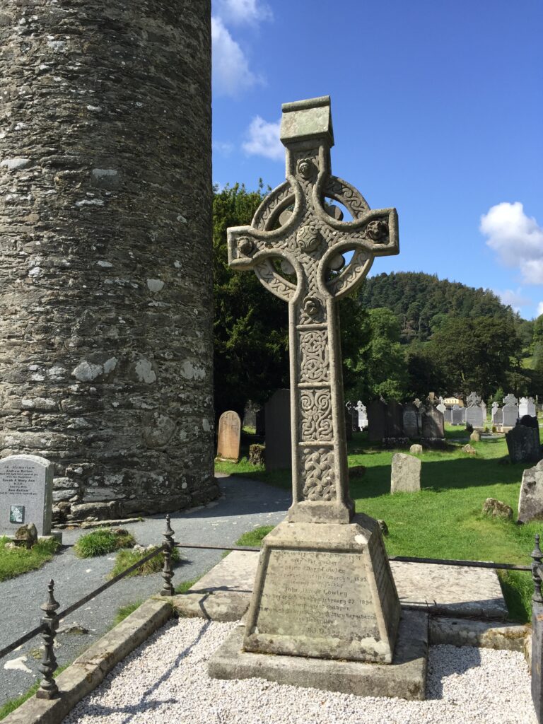 Large stone Celtic cross in cemetery surrounded by trees and grassland in Glendalough, Ireland